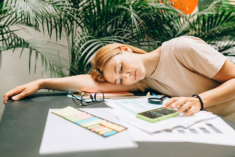 Woman lying on table with her glasses off trying to fill out her tax forms using her phone calculator